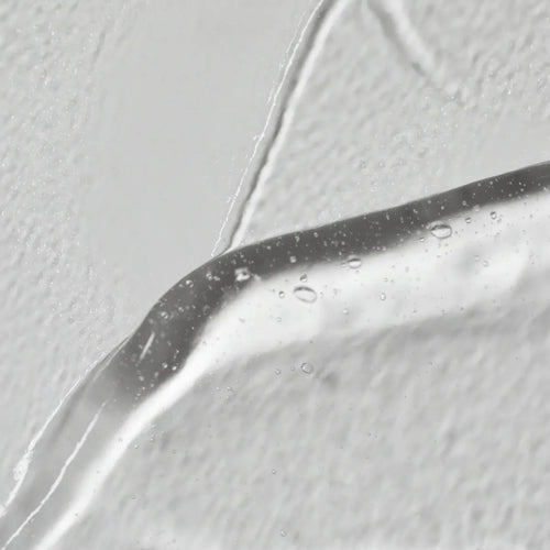 Close-up van Solawaves LightBoost Wand Activating Serum, een heldere gel met hyaluronzuur. Het bevat luchtbelletjes tegen een gestructureerde witte achtergrond, waardoor het gladde, glanzende oppervlak wordt benadrukt in contrast met de ruwe achtergrond.