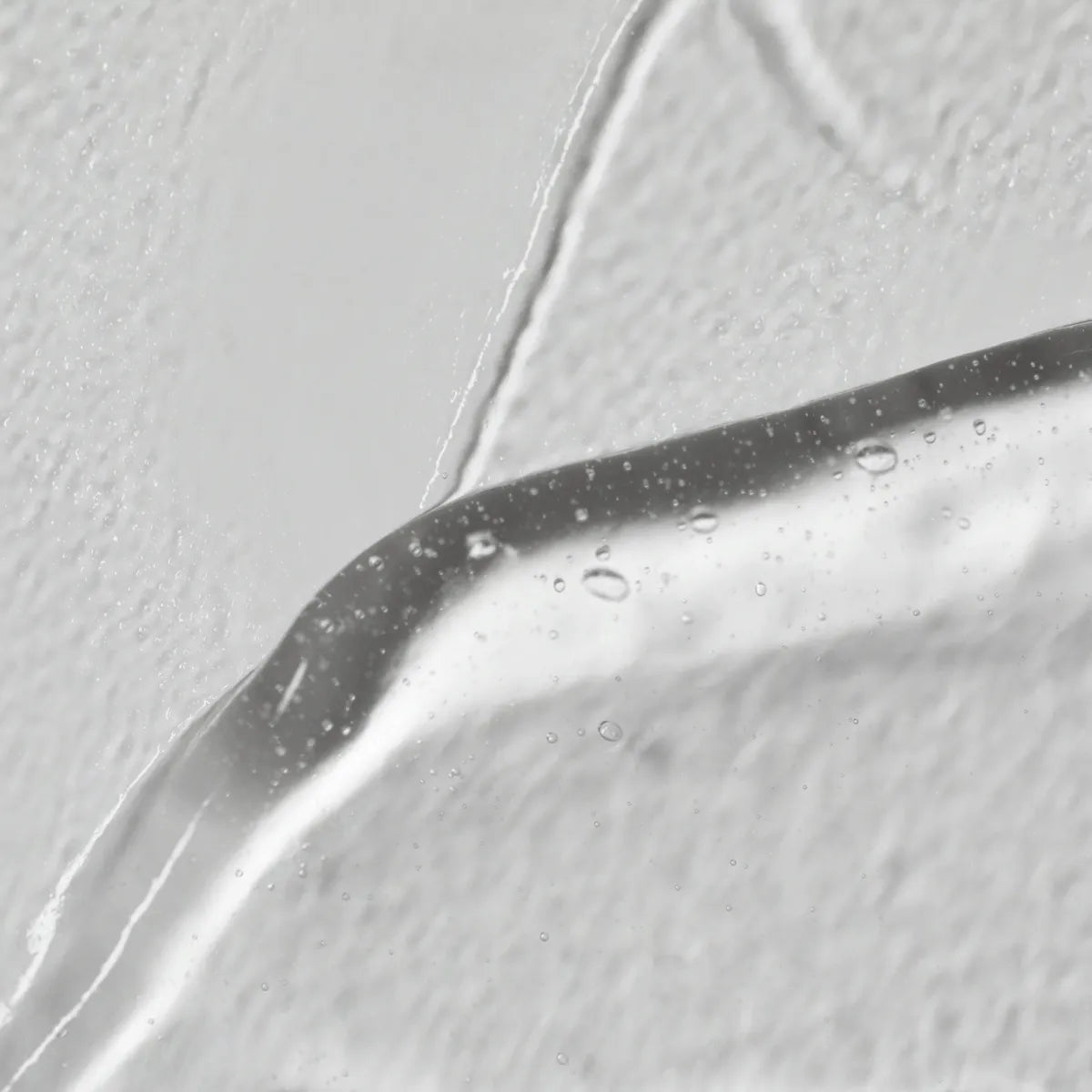Close-up van Solawaves LightBoost Wand Activating Serum, een heldere gel met hyaluronzuur. Het bevat luchtbelletjes tegen een gestructureerde witte achtergrond, waardoor het gladde, glanzende oppervlak wordt benadrukt in contrast met de ruwe achtergrond.